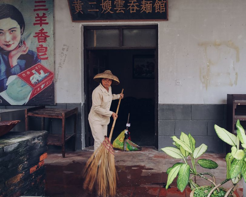 Asian woman in straw hat sweeping courtyard outside traditional building.